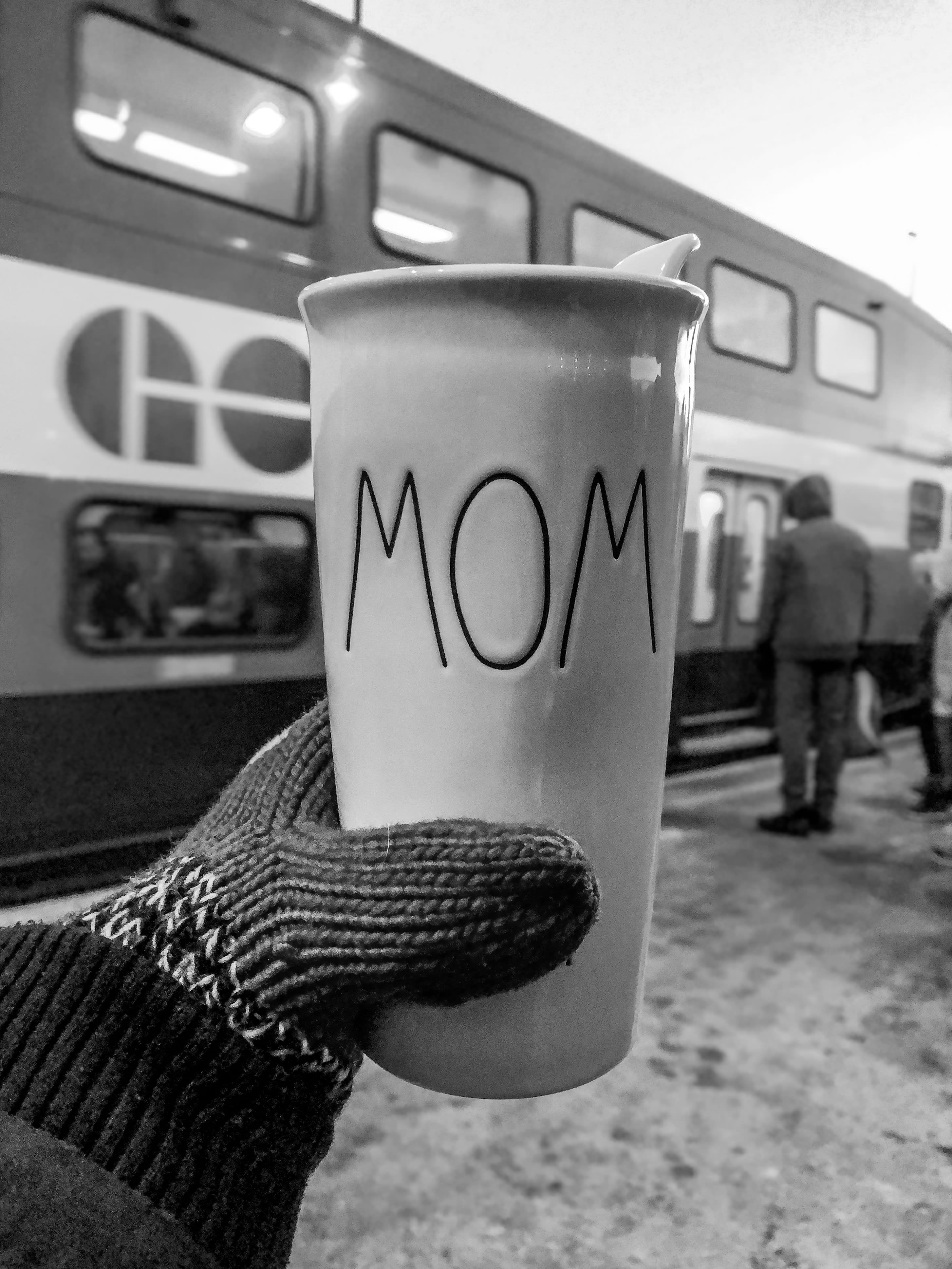 A mitted hand holds up a coffee cup with 'Mom' written on it