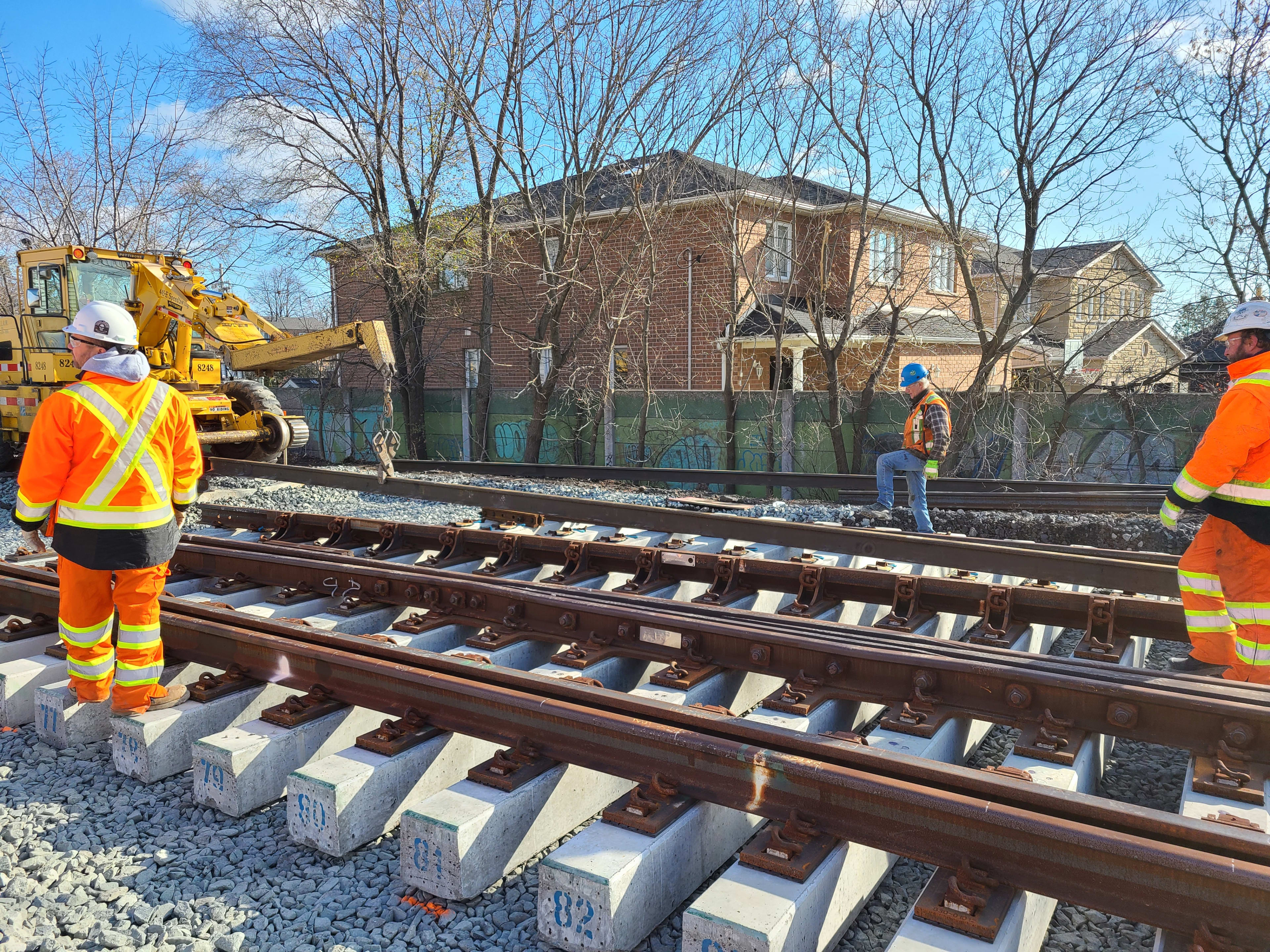 Workers stand next to new sections of rail with concrete rail ties as machinery works in the back...