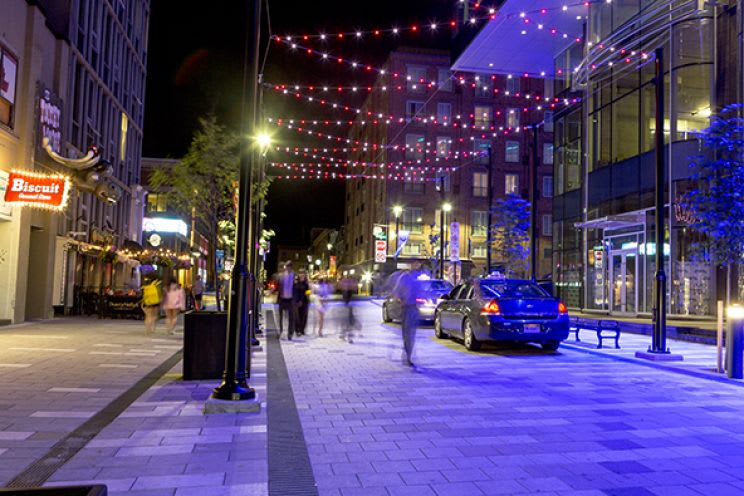 A street at night with festive, string lights and stylish streetscaping elements, including bench...