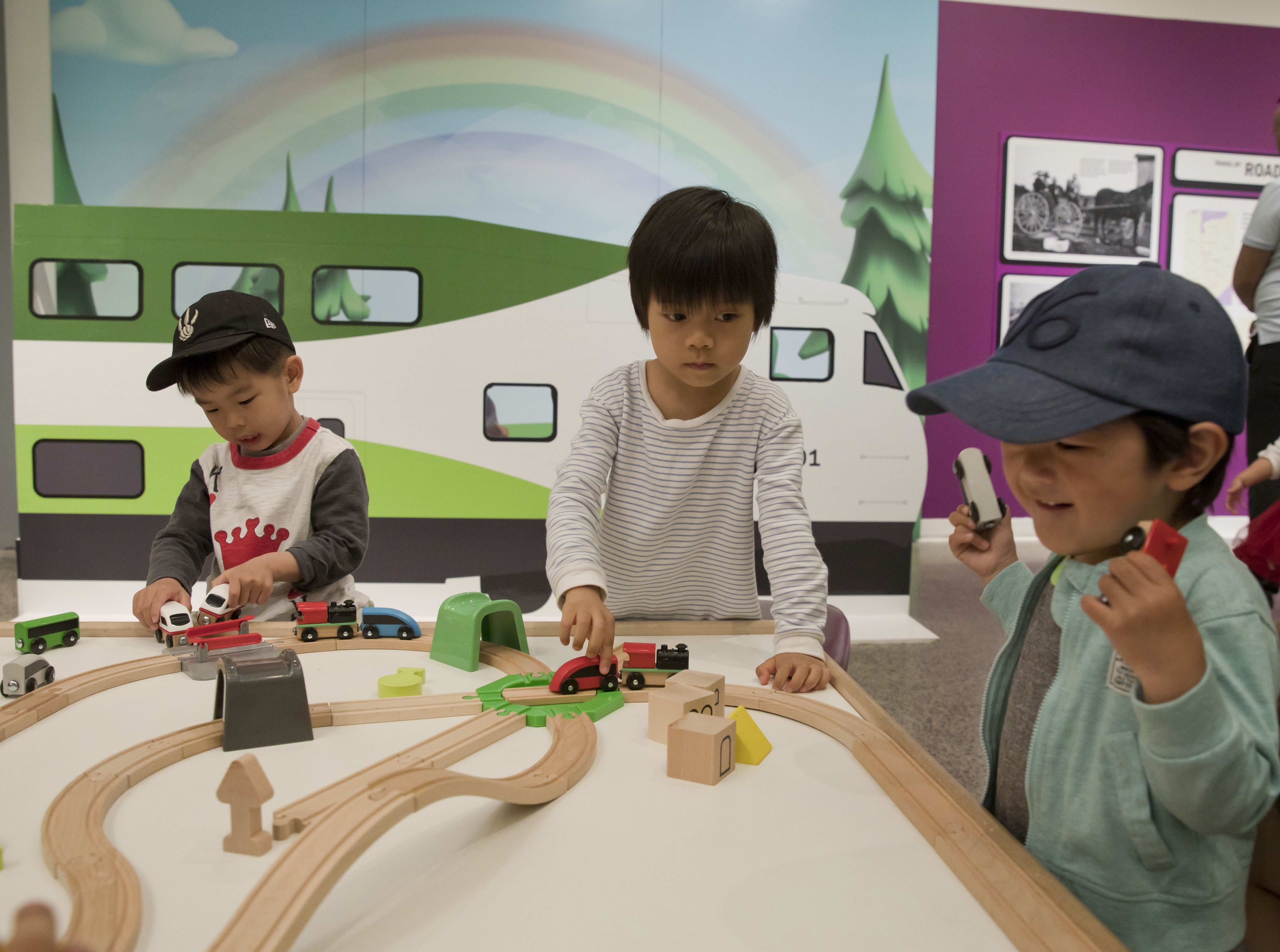 Three children play with cars and a wooden roadway on an activity table.