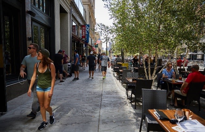 A wide sidewalk with a busy patio and vegetation separating pedestrians from the street.
