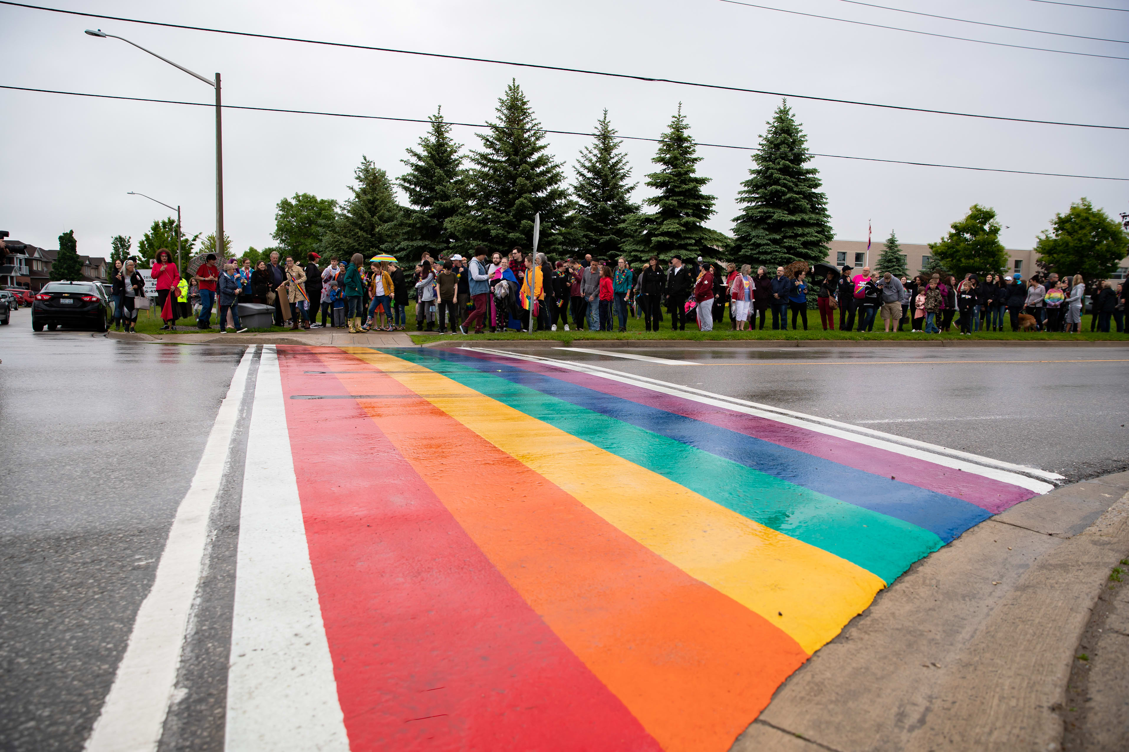 A crowd of people stand near a rainbow coloured crosswalk.