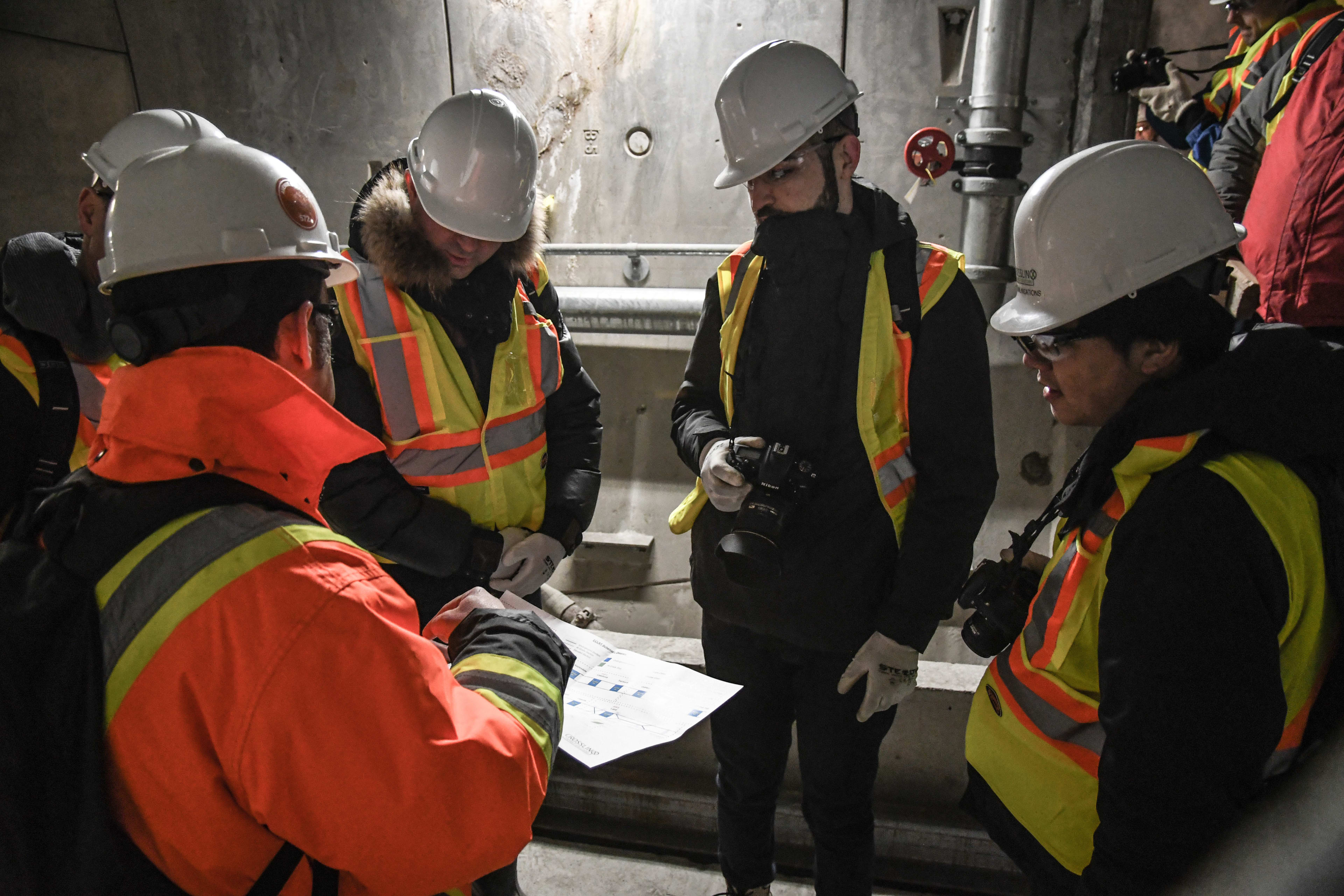Five people, all with hardhats and vests, look down at paperwork.