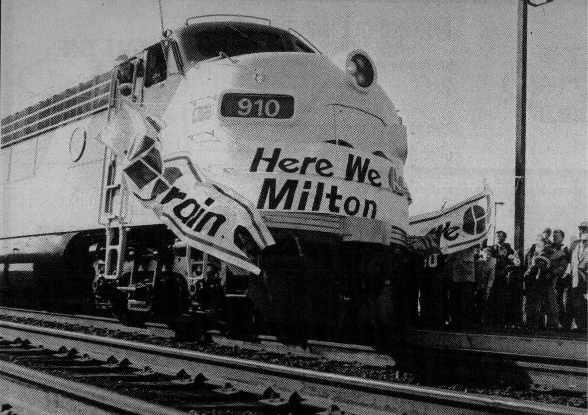 A train breaks a ribbon that says 'Here we GO Milton'.