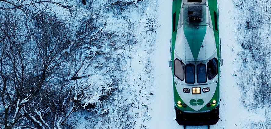 A GO train runs along a rail line, as snow sits arounds it.