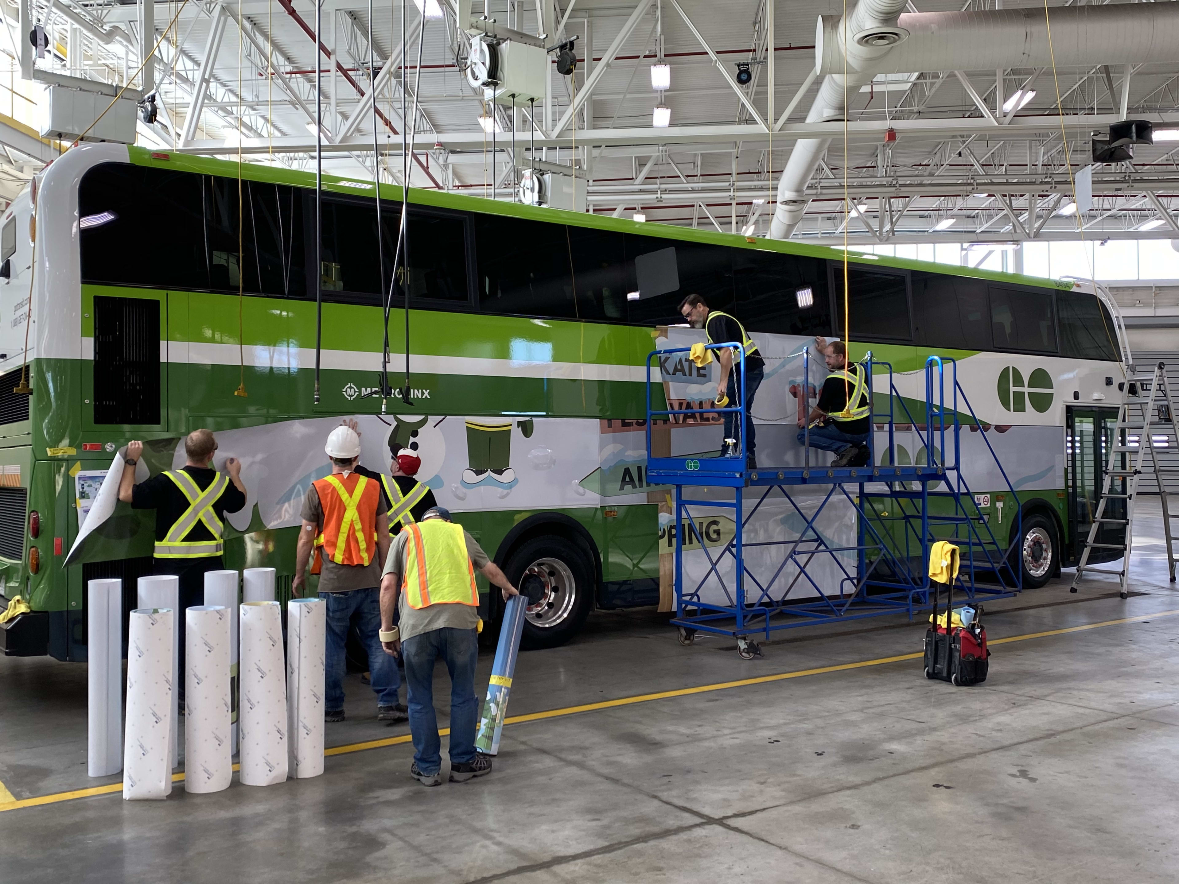 Crews stick vinyl panels onto the side of a GO bus.