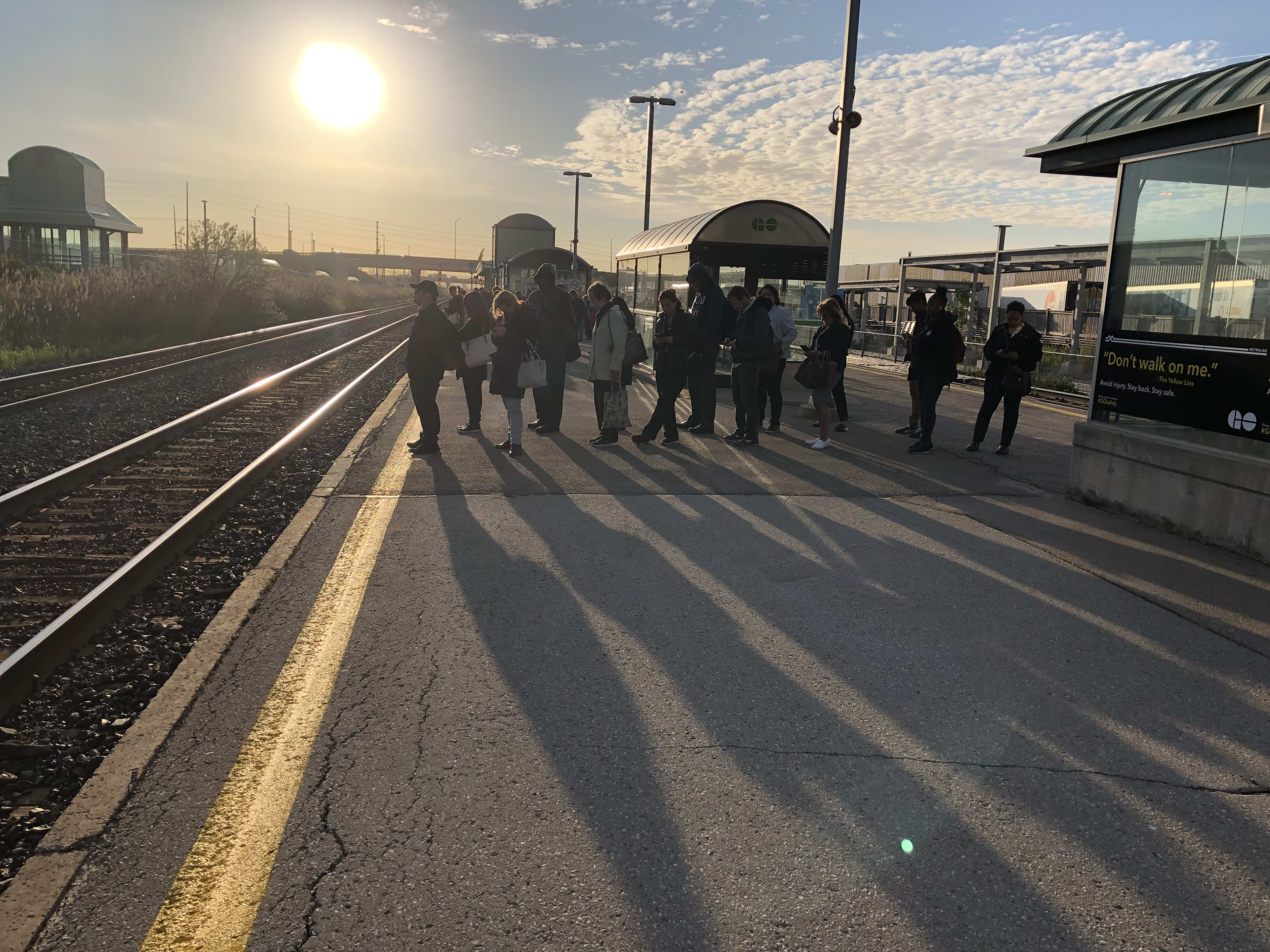 A line of customers produce long shadows on the platform, as an early day sun rises behind them.