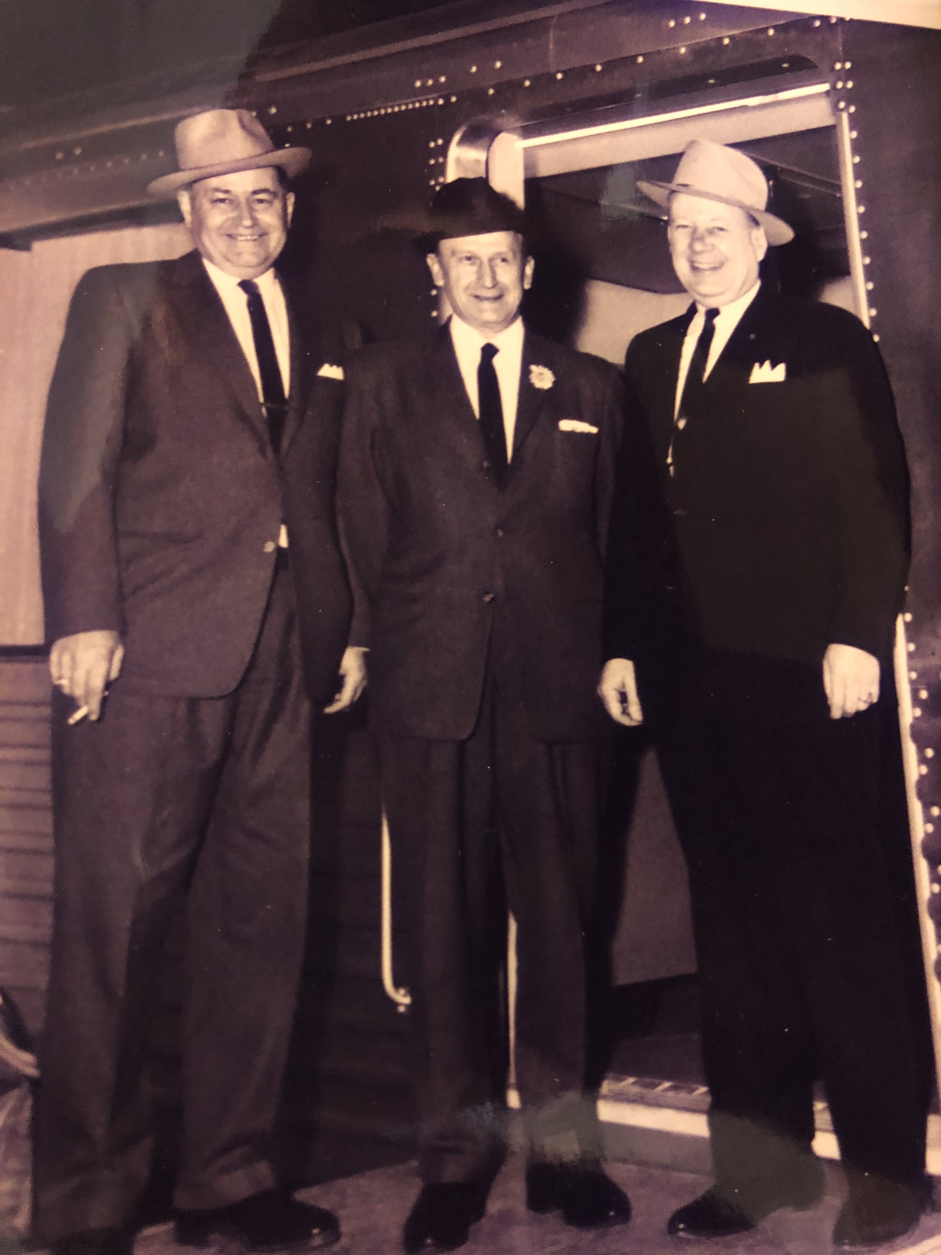 Three men in suits and hats pose in front of a train door.