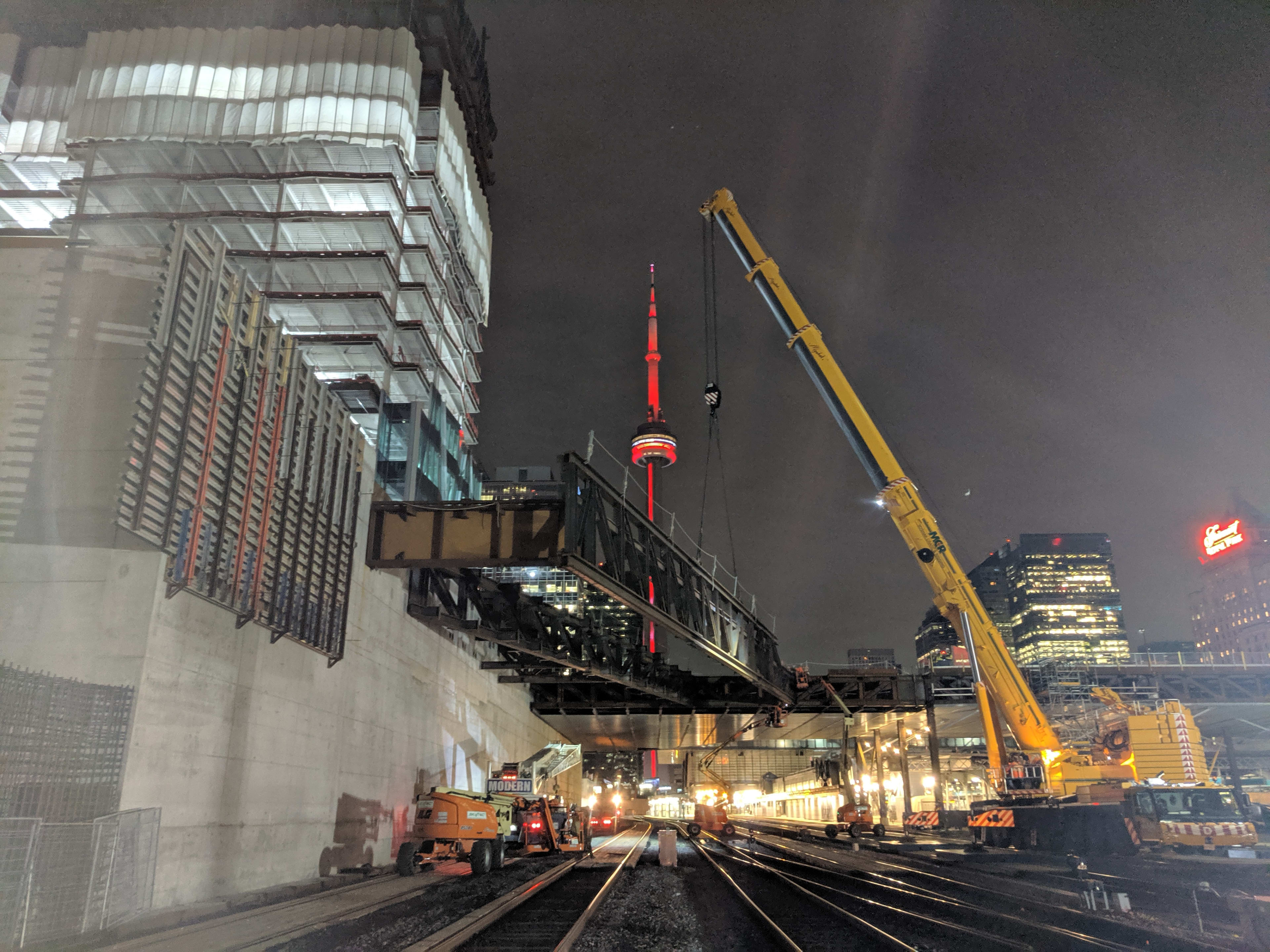 large yellow crane lifts big piece of metal into place with the lit up CN Tower in the backgound.