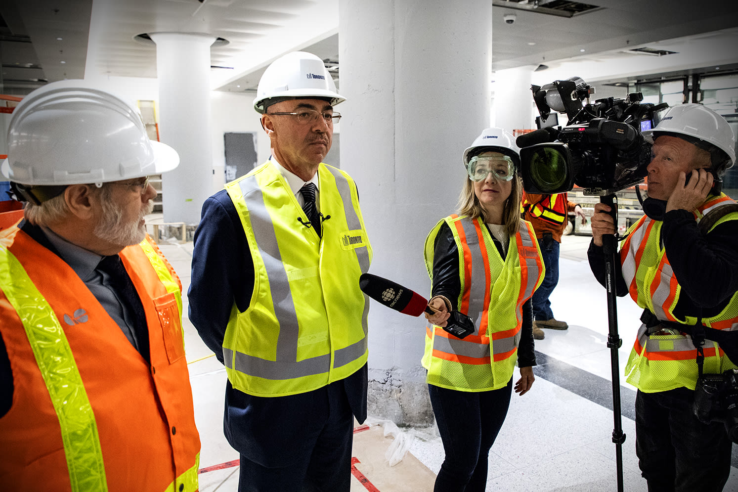 A cameraman and reporter interview two officials amid construction work.