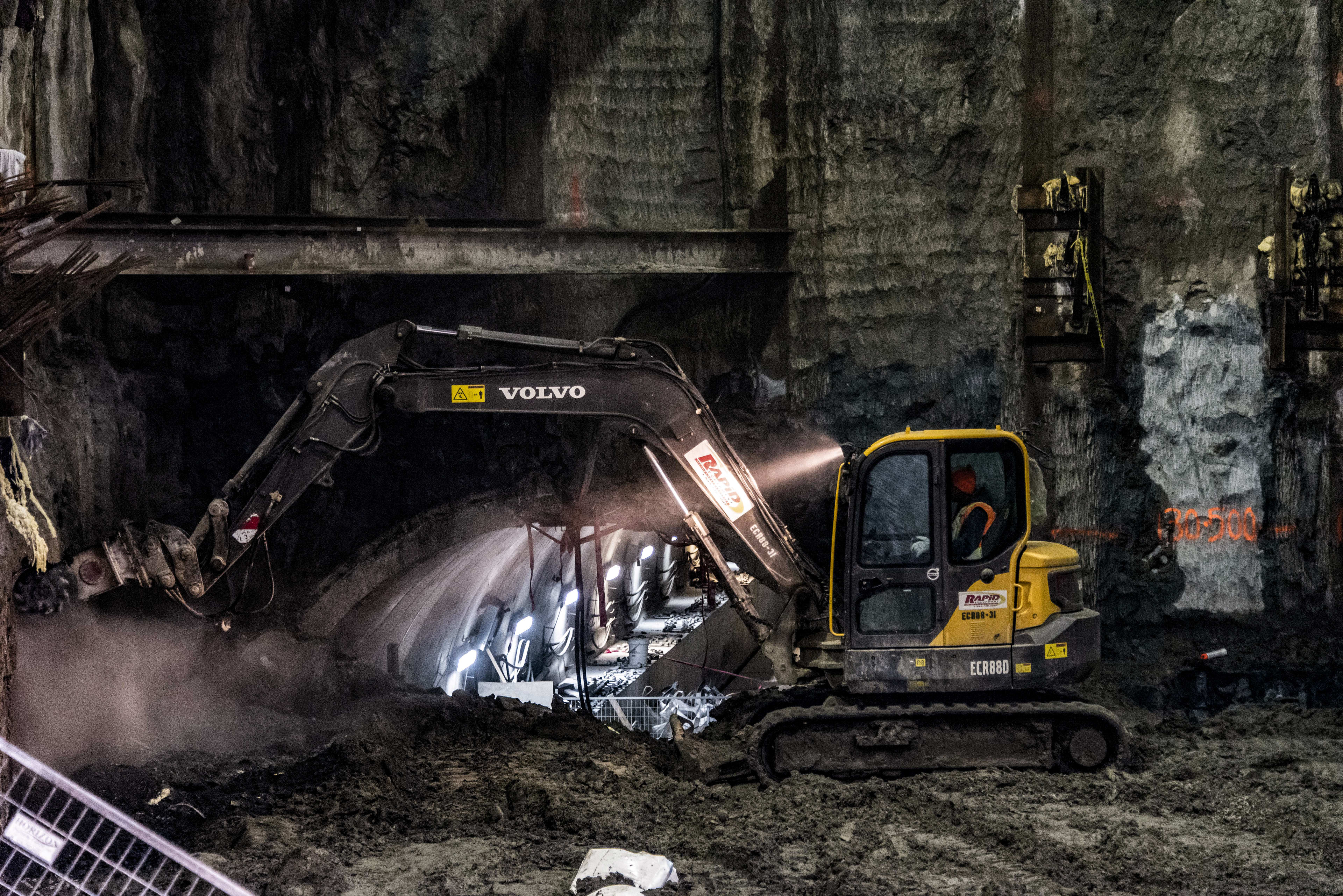 An earth mover digs into dirt, with a tunnel just behind.