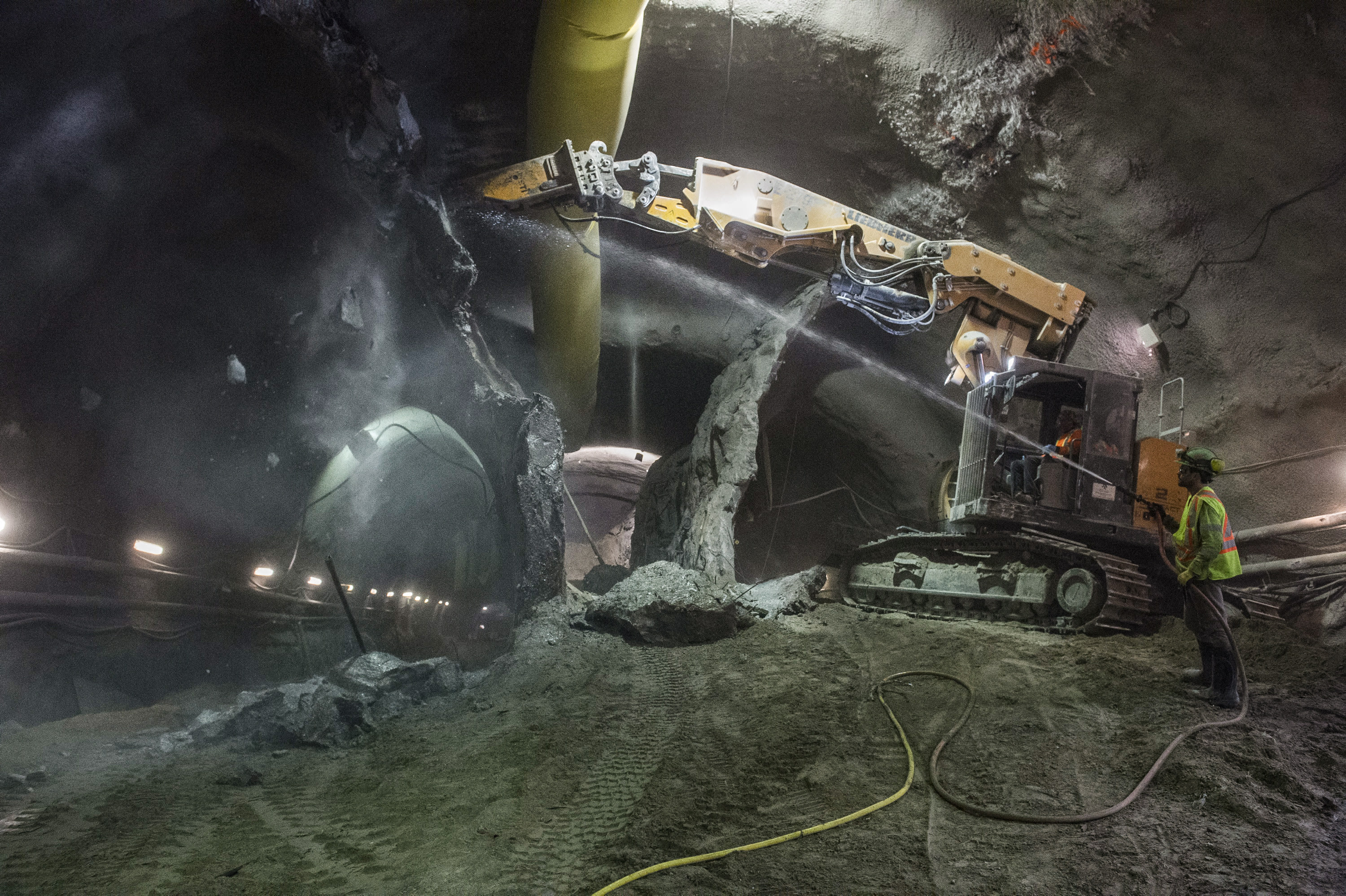 A crew member and digger chip away at rock and ground in a tunnel.