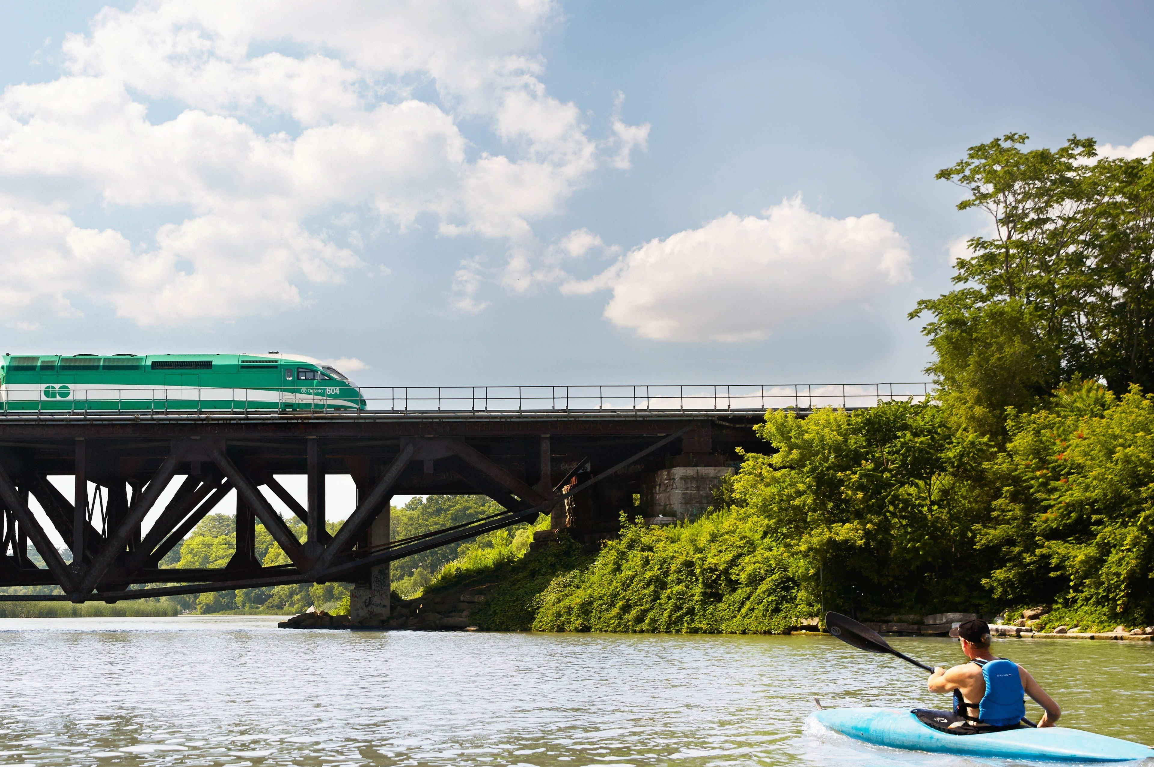 A GO train crosses over a bridge as man paddles a kyack on a waterway below.