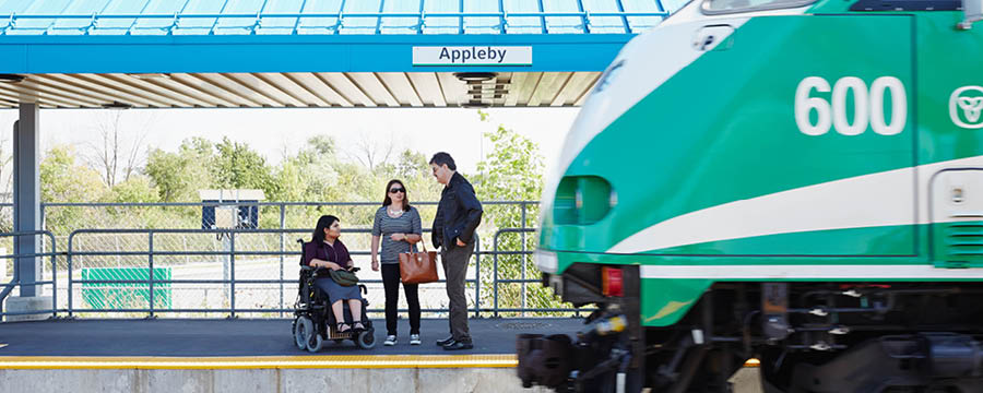Femme en fauteuil roulant avec deux autres personnes debout sur un quai et un train GO arrivant.