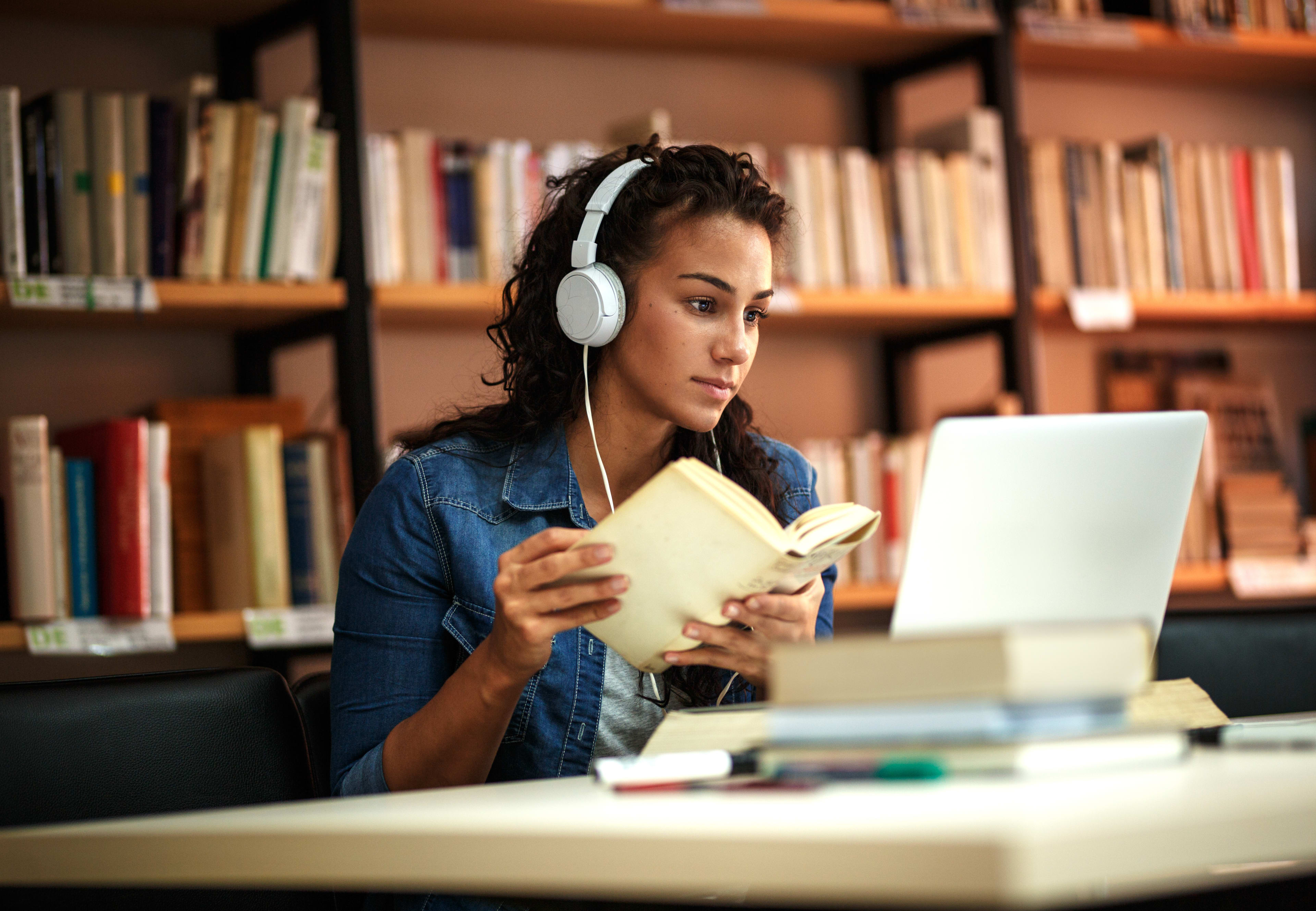 Person at the library studying on laptop with headphones and book in hand