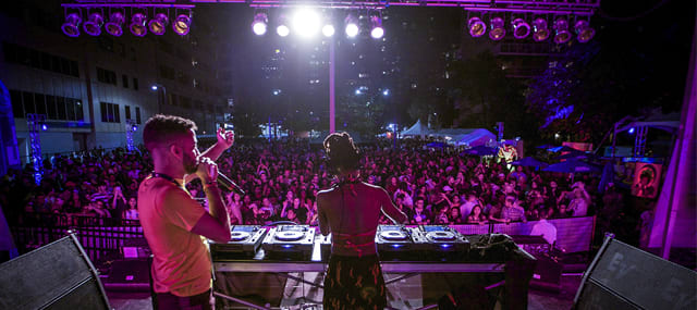 DJ onstage at a nightclub playing a mixer with announcer to the left on stage in front of big crowd