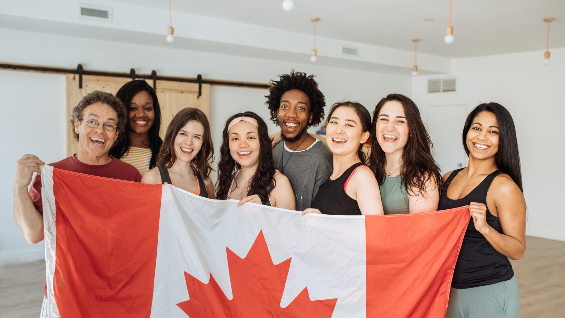 A group of 8 people of various genders and backgrounds holding up a Canadian flag