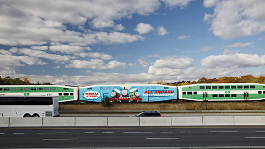 A train rides along a highway, with a special Thomas the Tank car